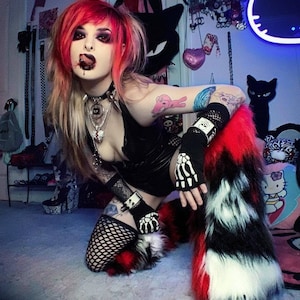 May include: A woman with red and blonde hair is wearing a black outfit with a silver necklace and a skull-shaped ring. She is wearing black fishnet stockings and red and white furry leg warmers. She has a tattoo on her arm and is holding a black and white skeleton-shaped glove.