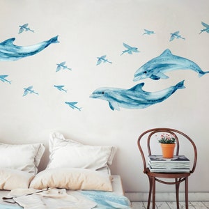 May include: A white wall with blue watercolor dolphin and flying fish decals. The decals are arranged in a playful pattern, creating a whimsical underwater scene.