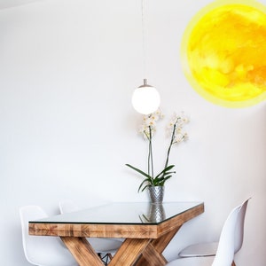 Puede incluir: Una mesa de comedor de madera con una tapa de vidrio y una base en forma de X pintada de blanco. Cuatro sillas blancas con patas de madera rodean la mesa. Una planta de orquídea en maceta se encuentra sobre la mesa. Un gráfico de sol amarillo está en la pared detrás de la mesa.