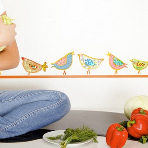 May include: A woman in a white shirt and blue jeans holds a bunch of red bell peppers and a head of cabbage in her hands. The kitchen counter is decorated with a row of colorful bird stickers.