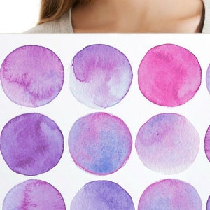 Könnte beinhalten: Eine weiße Leinwand mit 15 Aquarellkreisen in Violett-, Rosa- und Blautönen. Die Kreise sind in drei Reihen zu fünf angeordnet. Die Leinwand wird von den Händen einer Person gehalten.