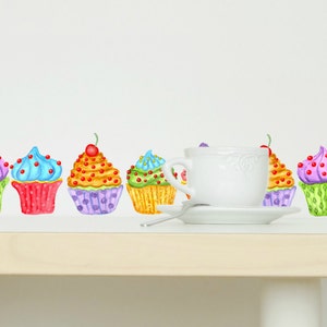 May include: A row of colorful cupcakes with white frosting and sprinkles, a white teacup and saucer on a light wood table with a white chair in the background.
