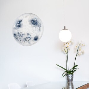 Puede incluir: Una mesa de comedor de madera con una tapa de vidrio y un acabado pintado de blanco. La mesa tiene una base en forma de cruz y está rodeada de cuatro sillas blancas. Una lámpara colgante blanca cuelga sobre la mesa, y una planta en maceta con flores blancas se coloca sobre la mesa.