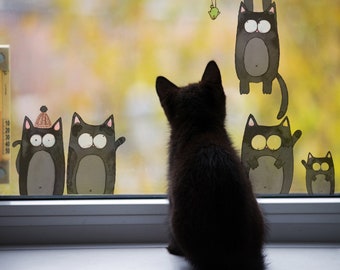May include: A black cat sits on a windowsill looking out at a window with cartoon cats on it. The cartoon cats are black and white and have different expressions. One cartoon cat is wearing a hat.