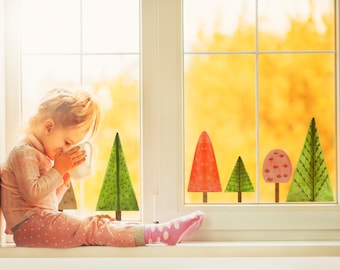 May include: A young girl sits by a window with a cup in her hands.  The window has five colourful tree stickers on it.  The trees are green, red, and pink.