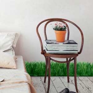 May include: A wooden chair with a stack of books and a potted plant with pink flowers on the seat. The chair is sitting on a white floor with a green grass border.