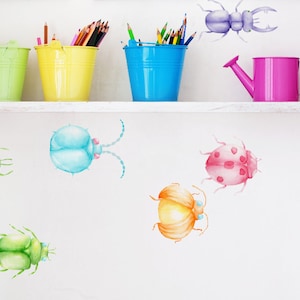 Könnte beinhalten: Ein weißes Regal mit drei bunten Eimern gefüllt mit Buntstiften, einer rosa Gießkanne und einem lila Käfer-Wandaufkleber. Auf der weißen Wand unter dem Regal befinden sich auch mehrere bunte Käfer-Wandaufkleber.