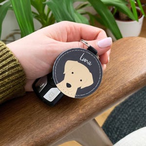 May include: A circular, dark grey keyring featuring a light tan dog illustration and the name "Luna" in white script. The keyring is attached to a black strap and silver ring. A car key fob is visible.