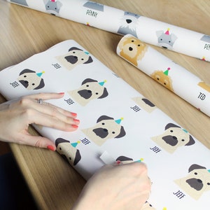 K&ouml;nnte beinhalten: Wei&szlig;es Geschenkpapier mit einem sich wiederholenden Muster von Mops, die Partyh&uuml;te tragen. Die M&ouml;pse sind beige mit schwarzer Nase und Augen. Der Text "Jeff" ist unter jedem Mops gedruckt. Eine Hand h&auml;lt das Papier und ein St&uuml;ck Klebeband.