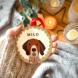 May include: A round, decorative ornament featuring a dog illustration with the name "MILO" in black text. The dog has brown and white markings. The ornament has a scalloped edge and a red ribbon for hanging.