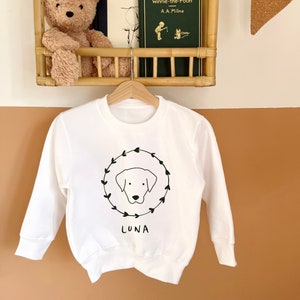 May include: White sweatshirt with a black and white illustration of a dog's head inside a wreath. The text "LUNA" is printed below the illustration.