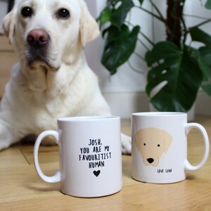 May include: Two white ceramic mugs with black text and a heart. One mug says "Josh, you are my favouritist human" with a black heart. The other mug has a drawing of a yellow labrador and says "Love Luna". A yellow labrador dog is sitting in the foreground.