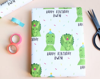 May include: White wrapping paper with a green dinosaur pattern. The dinosaurs are wearing party hats and there are colorful confetti scattered across the paper. The text "Happy Birthday Owen!" is repeated throughout the paper.