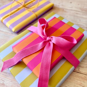 May include: Two wrapped gifts with colorful striped paper. One is wrapped in yellow and purple stripes and tied with twine. The other is wrapped in orange and pink stripes and tied with a pink ribbon. A third gift is visible with yellow and light blue stripes.