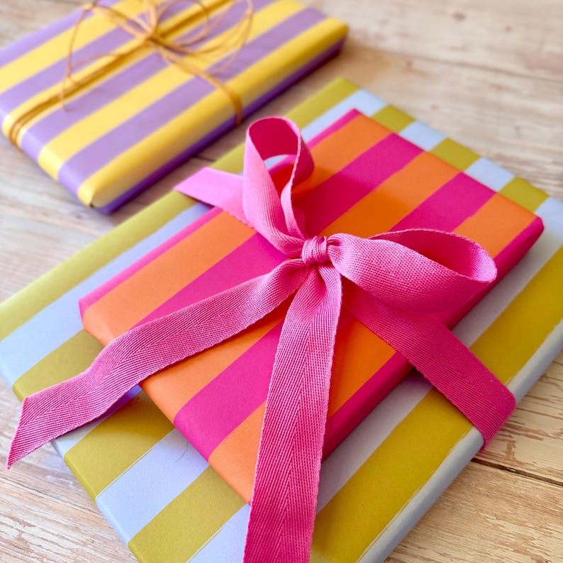 May include: Two wrapped gifts with colorful striped paper. One is wrapped in yellow and purple stripes and tied with twine. The other is wrapped in orange and pink stripes and tied with a pink ribbon. A third gift is visible with yellow and light blue stripes.