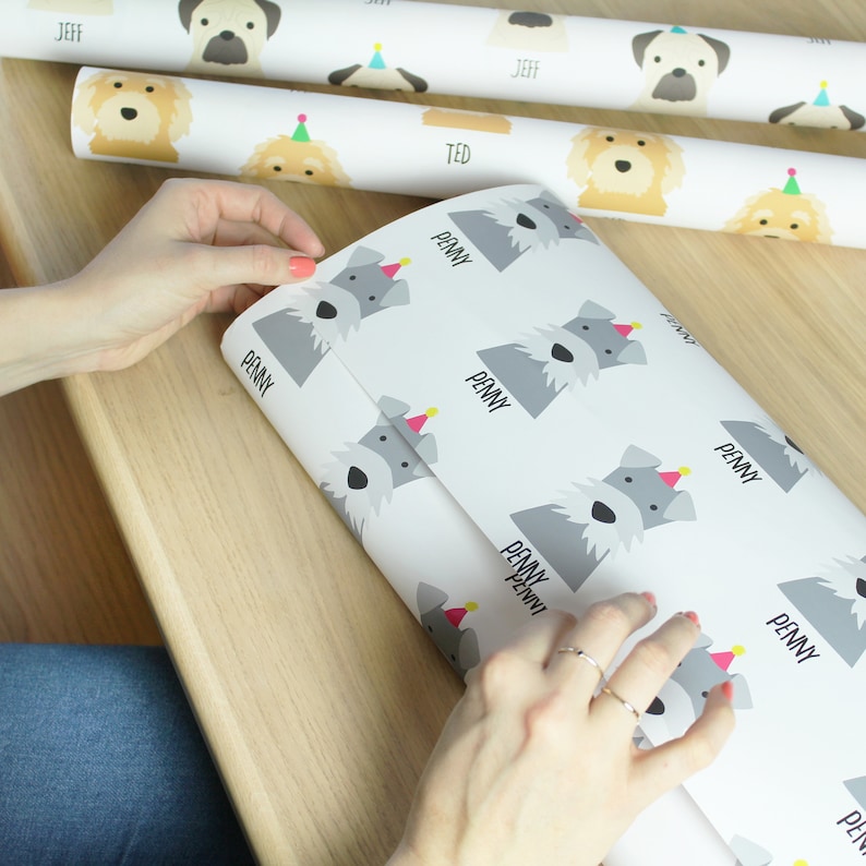K&ouml;nnte beinhalten: Wei&szlig;es Geschenkpapier mit einem sich wiederholenden Muster aus grauen Schnauzer-Hunden, die rosa Partyh&uuml;te tragen. Das Papier tr&auml;gt den Namen "Penny" aufgedruckt.