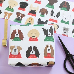 May include: White wrapping paper with a repeating pattern of dogs wearing Christmas sweaters. The dogs are all different breeds and have different expressions. The sweaters are red and green with the text "Merry Christmas" on them.
