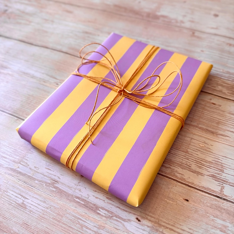 May include: A wrapped gift with a striped pattern of yellow and purple. The gift is tied with a brown twine bow. The gift is on a light brown wooden surface.