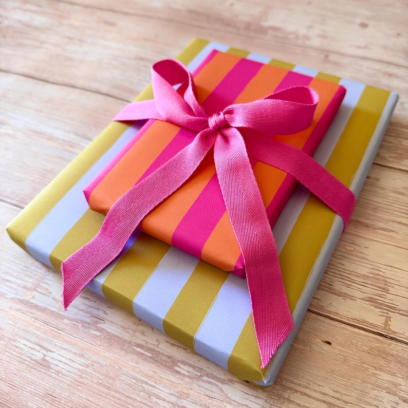 May include: Two wrapped gifts with colorful striped paper. The top gift has pink and orange stripes, and the bottom gift has yellow and white stripes. A bright pink ribbon is tied around both gifts.