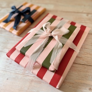 May include: Two rectangular gifts wrapped in striped paper. The larger gift features vertical pink and dark red stripes tied with a frayed light pink ribbon. The smaller gift in the background has orange and pink stripes with a dark blue ribbon.