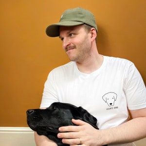 Könnte beinhalten: Ein Mann trägt ein weißes T-Shirt mit dem Text "Fleur's Dad" und einer schwarz-weißen Illustration eines Hundes. Er hält einen schwarzen Labrador Retriever in seinen Armen. Er trägt eine grüne Baseballkappe.
