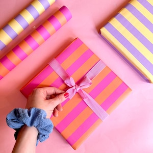 May include: A square gift wrapped in pink and orange striped paper, tied with a pink ribbon and bow. Rolls of striped wrapping paper in pink, orange, and purple are also visible. A blue scrunchie is on a person's wrist.