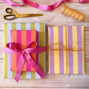 May include: Overhead shot of wrapped gifts with colorful striped paper. One gift is wrapped in pink, orange, and yellow striped paper and tied with a pink ribbon. Another is wrapped in yellow and purple striped paper and tied with twine. Gold scissors and pink ribbon are also visible.