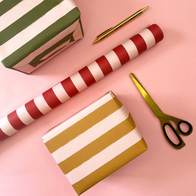 May include: Overhead shot of gift wrapping supplies on a pink surface. Includes wrapped boxes with green and gold stripes, a roll of red and white striped wrapping paper, gold scissors, and a gold pen. The items are arranged diagonally.
