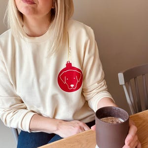 Könnte beinhalten: Ein cremefarbener Pullover mit einem roten Weihnachtsbaumschmuck, der einen Hundekopf zeigt. Der Schmuck hängt an einer goldenen Schnur mit dem Wort "Luminous" darauf.