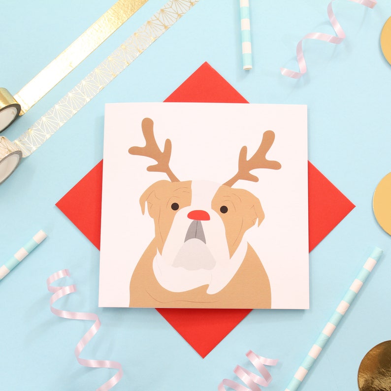 May include: A white card with a brown and white bulldog wearing a red reindeer antler headband. The card is on a red background with gold and blue accents.