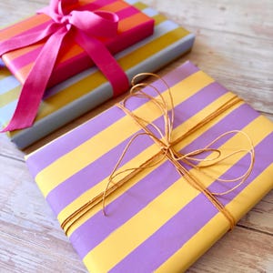 May include: Two wrapped gifts with colorful striped paper. One is wrapped in purple and yellow stripes and tied with twine. The other is wrapped in pink and orange stripes and tied with a pink ribbon.