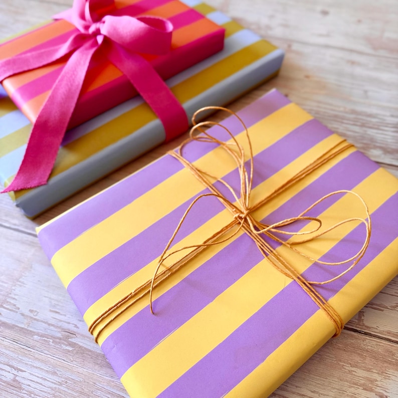 May include: Two wrapped gifts with colorful striped paper. One is wrapped in purple and yellow stripes and tied with twine. The other is wrapped in pink and orange stripes and tied with a pink ribbon.