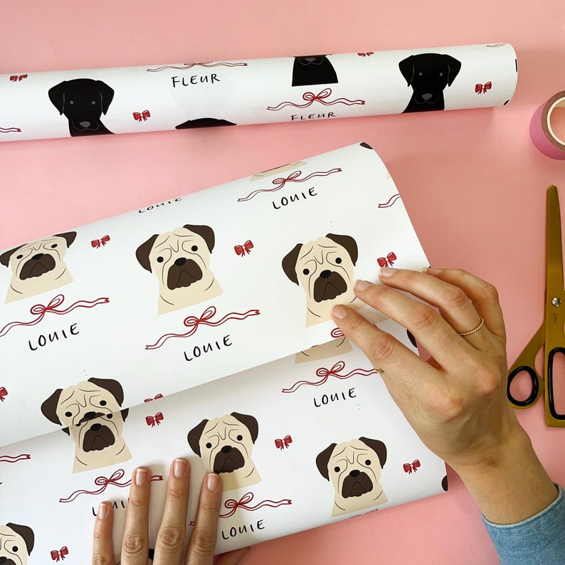 May include: White wrapping paper with a repeating pattern of pugs and red bows. The paper has the name "Louie" printed on it.