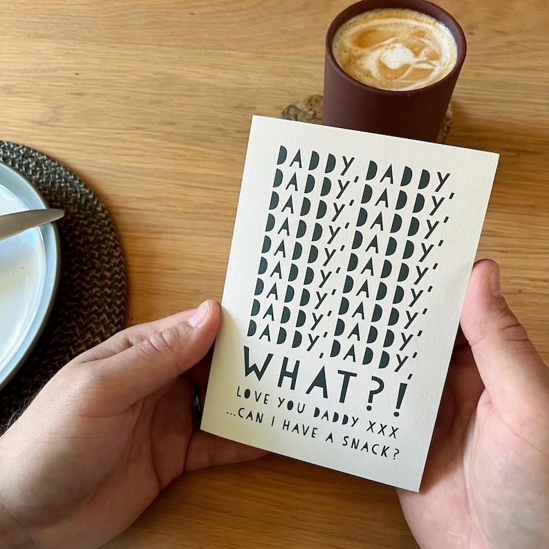 May include: A greeting card with the words "DADDY, DADDY" repeated multiple times, followed by "WHAT?!" and "LOVE YOU DADDY XXX...CAN I HAVE A SNACK?" in black text. The card is held in front of a brown mug with a coffee drink.