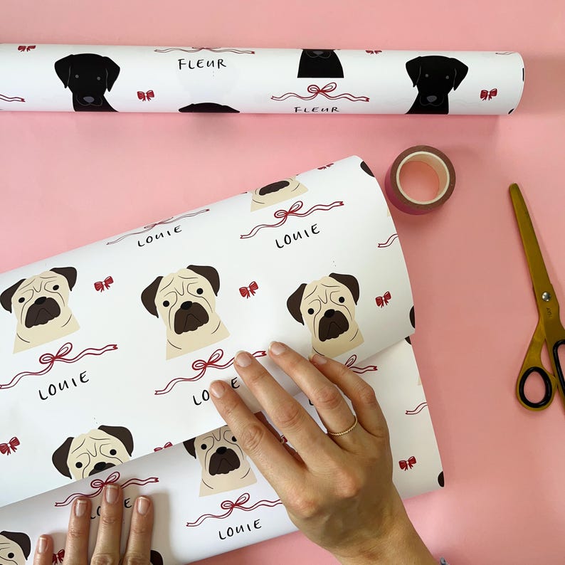 May include: White wrapping paper with a repeating pattern of pug dog faces and red bows. The paper is labeled "Louie" in black handwriting. A roll of wrapping paper with black Labrador Retriever faces and red bows is also visible in the background. The paper is labeled "Fleur" in black handwriting.