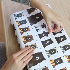 May include: White wrapping paper with a repeating pattern of black bears wearing different colored scarves. The bears are wearing scarves in the colors of the Hogwarts houses from the Harry Potter series.