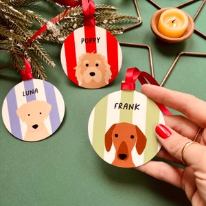 May include: Three round Christmas ornaments with dog illustrations and names: "POPPY" on red and white stripes, "LUNA" on blue and white stripes, and "FRANK" on green and white stripes. Each ornament has a red ribbon for hanging.