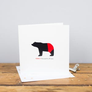May include: A white greeting card with a black bear wearing red shorts. The phrase "FANCY the pants off you" is printed in red beneath the bear. The card is presented on a wooden surface.