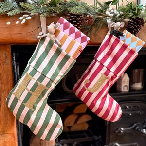 May include: Two fabric Christmas stockings hanging side by side. The left stocking features green and white vertical stripes with a gold glitter letter H. The right stocking has red and white vertical stripes with a gold glitter letter J.