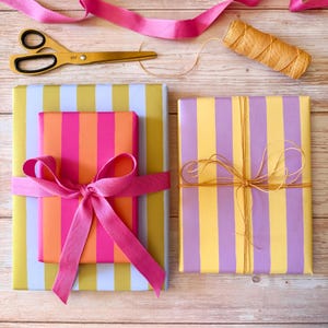 May include: Overhead shot of wrapped gifts with colorful striped paper. One gift is wrapped in pink, orange, and yellow stripes, tied with a pink ribbon. Another is wrapped in yellow and purple stripes, tied with twine. Gold scissors and pink ribbon are also visible.