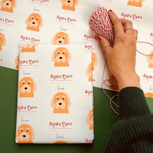 May include: White wrapping paper with a repeating pattern of a brown and white dog with the text "Santa Paws" and "Luna" in red. The paper is being wrapped around a gift box with red and white twine.