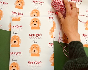 May include: White wrapping paper with a repeating pattern of a brown and white dog with the text "Santa Paws" and "Luna" in red. The paper is being wrapped around a gift box with red and white twine.