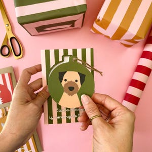 May include: A green and white striped card with a round, green ornament featuring a brown and white dog. The ornament has a brown string attached to it. The text on the card reads "Santa paws is coming to town".