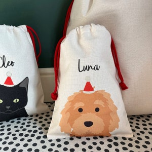 May include: Two personalised Christmas gift bags with red drawstrings. One bag features a black cat wearing a Santa hat and the name "Cleo" printed on the front. The other bag features a brown dog wearing a Santa hat and the name "Luna" printed on the front.