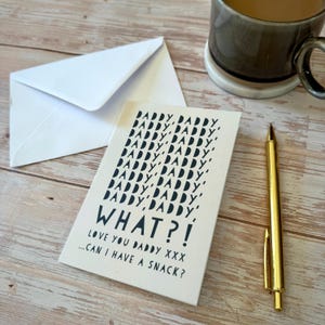 May include: A greeting card with the words "DADDY, DADDY, WHAT?!" in black text. The card is on a wooden surface, next to a white envelope and a gold pen. The card also says "LOVE YOU DADDY XXX...CAN I HAVE A SNACK?"