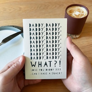 May include: A greeting card with the words "DADDY, DADDY, WHAT?!" in black font. The card is held in a person's hands. The text "LOVE YOU DADDY XXX...CAN I HAVE A SNACK?" is also on the card. A cup of coffee is in the background.
