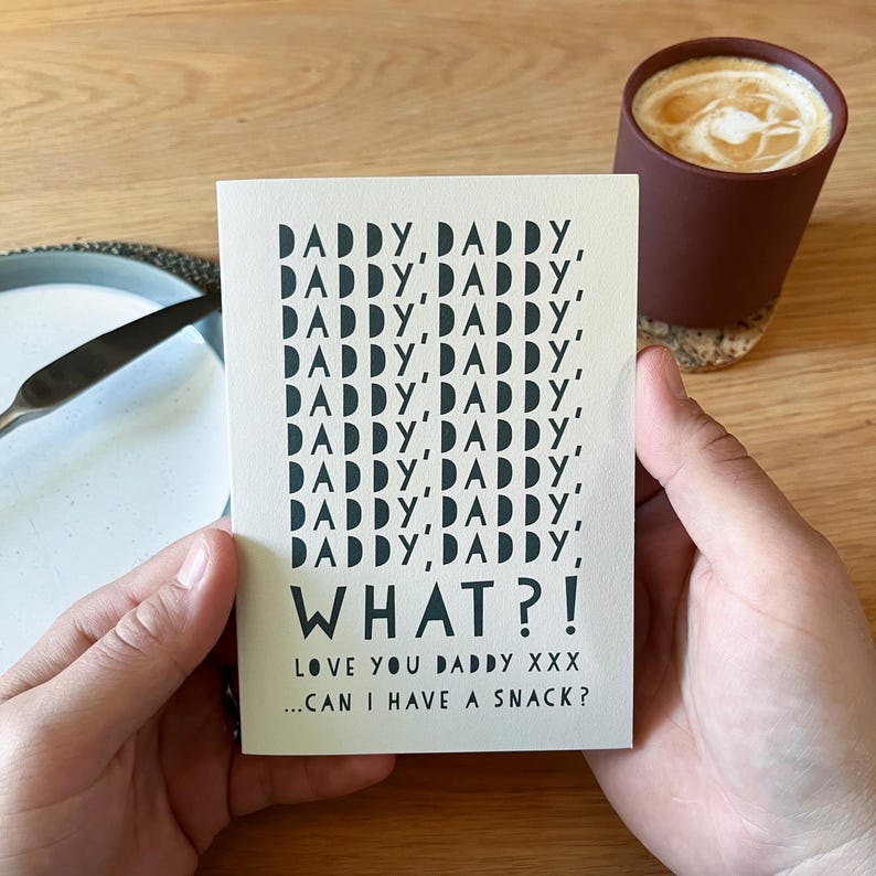 May include: A greeting card with the words "DADDY, DADDY, WHAT?!" in black font. The card is held in a person's hands. The text "LOVE YOU DADDY XXX...CAN I HAVE A SNACK?" is also on the card. A cup of coffee is in the background.