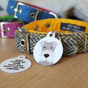May include: A brown, green and yellow tweed dog collar with a silver buckle and a white round dog tag. The dog tag has a black and white illustration of a small dog with a grey nose and a black nose. The dog tag also has text that reads "Fleur, 2 York Rd, York, YO19 6QQ, 07777 222 333, Chipped".