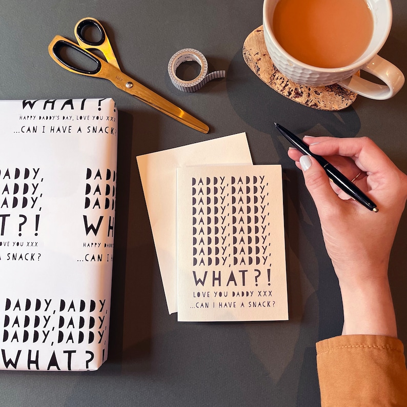 May include: A gift set with a greeting card and wrapping paper, both featuring the text "Daddy, Daddy, What?! Love You Daddy... Can I Have a Snack?" in black on a white background. Gold scissors and a roll of tape are also present.