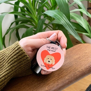 May include: A pink circular keyring featuring a dog illustration within a red heart. The text reads "THE BEST dog mum!" and "Love, Teddyx". The keyring has a silver ring and a black strap, perfect for keys or bags.